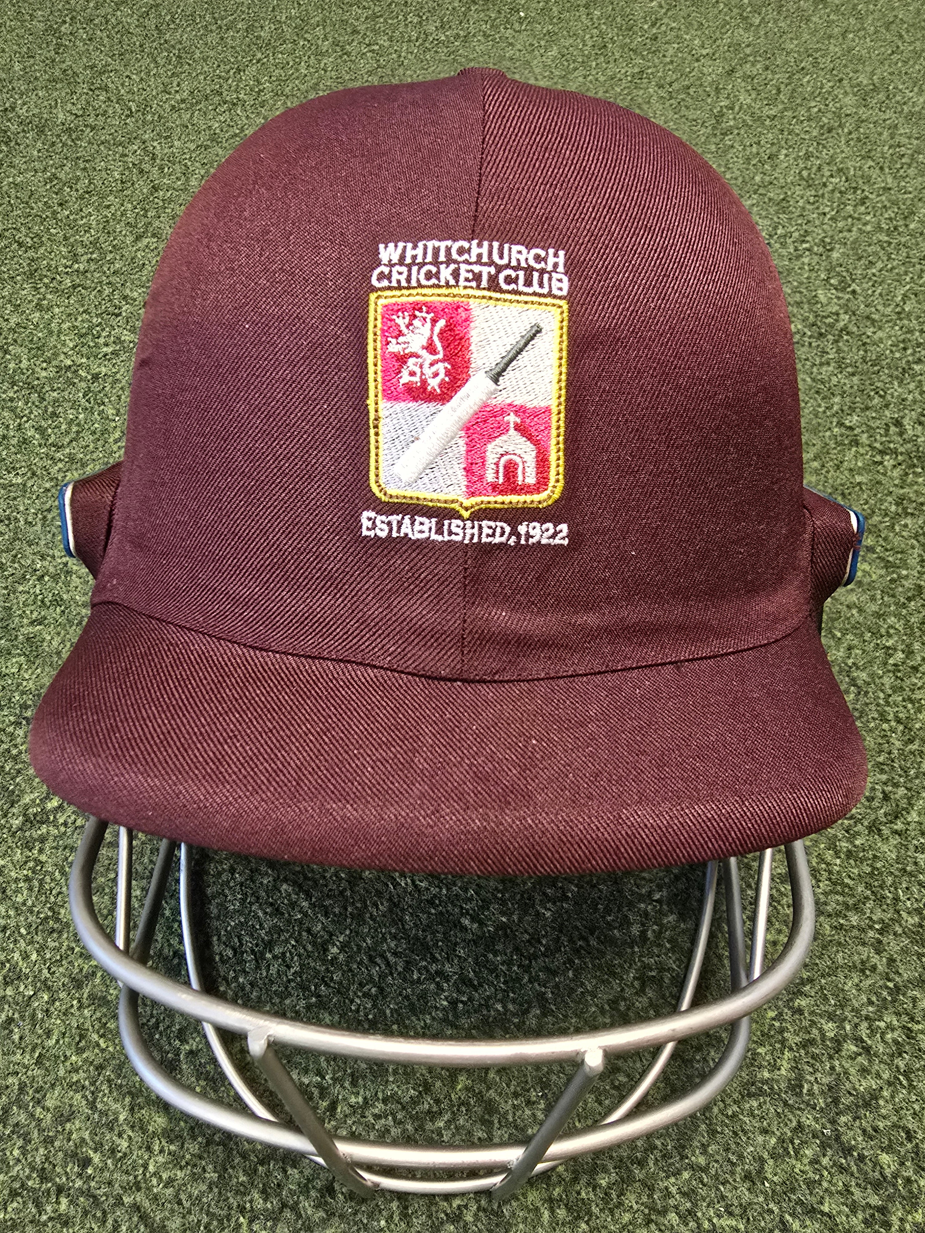 A maroon Custom Shrey Cricket Helmet with the Whitchurch Cricket Club logo—lion, bat, ball, and "Established 1922"—embroidered on the front sits on green turf.