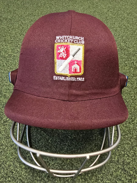 A maroon Custom Shrey Cricket Helmet with the Whitchurch Cricket Club logo—lion, bat, ball, and "Established 1922"—embroidered on the front sits on green turf.