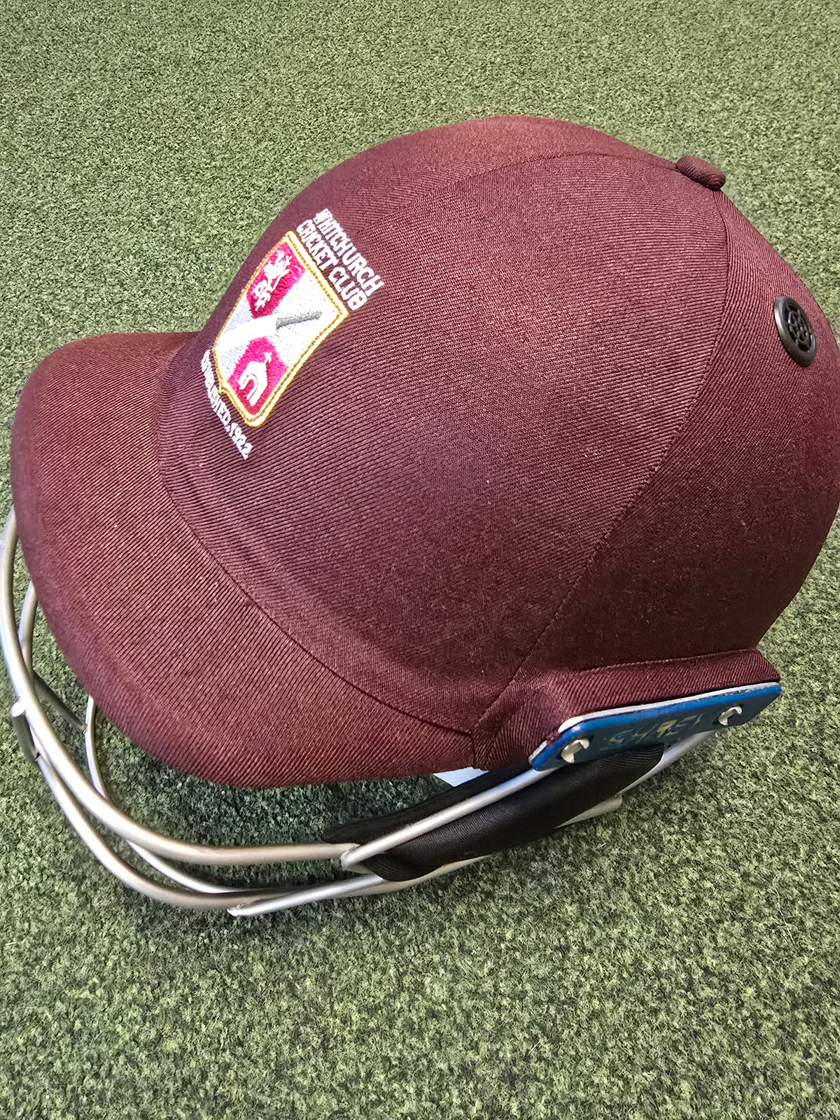 A Custom Shrey Cricket Helmet with the Northamptonshire County Cricket Club logo and metal face guard rests on a green carpet.