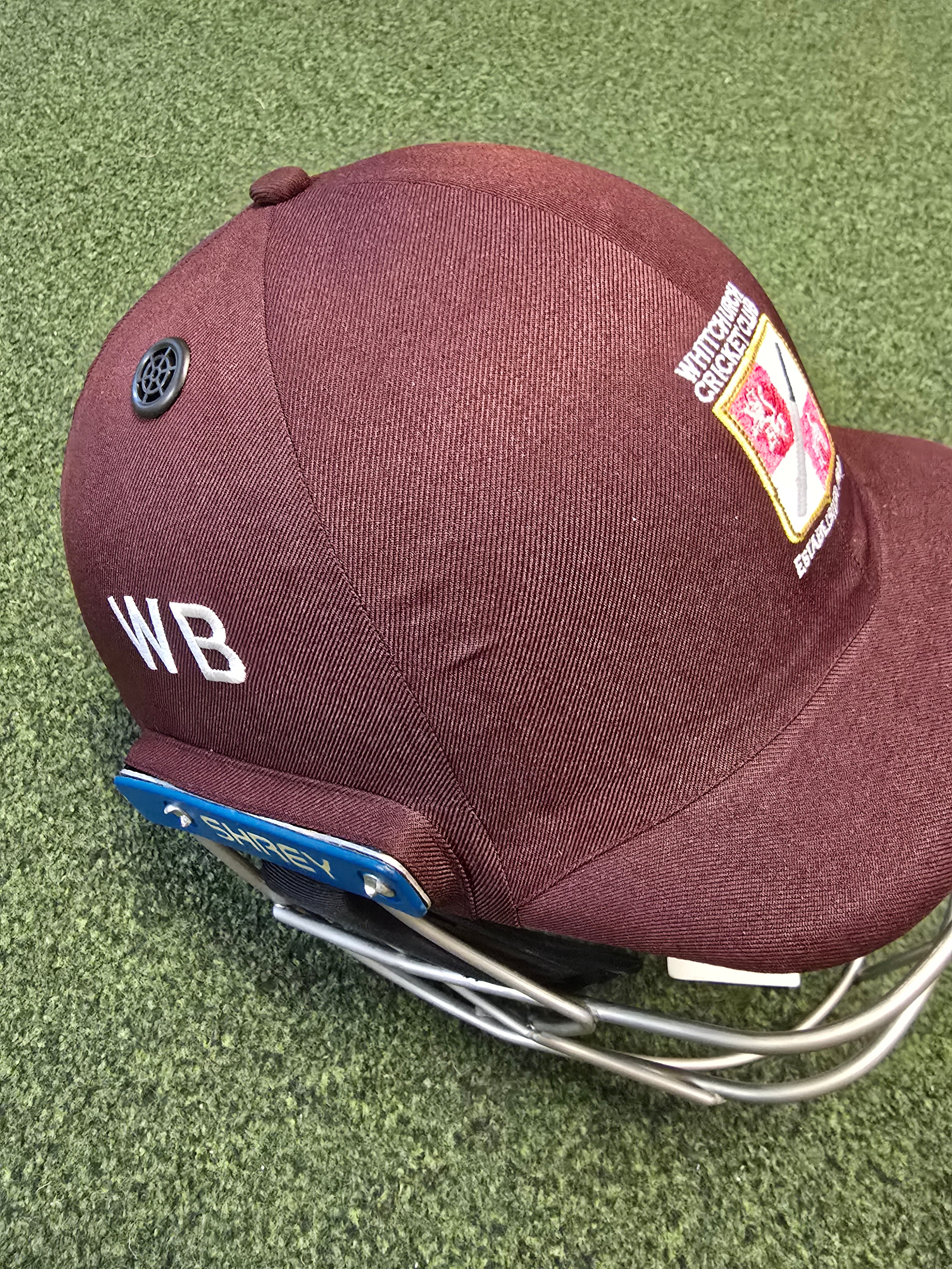 A maroon Custom Shrey Cricket Helmet by Shrey, featuring embroidered WB initials, the Northamptonshire County Cricket Club logo, and a blue tag, rests on green artificial turf.