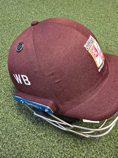 A maroon Custom Shrey Cricket Helmet by Shrey, featuring embroidered WB initials, the Northamptonshire County Cricket Club logo, and a blue tag, rests on green artificial turf.
