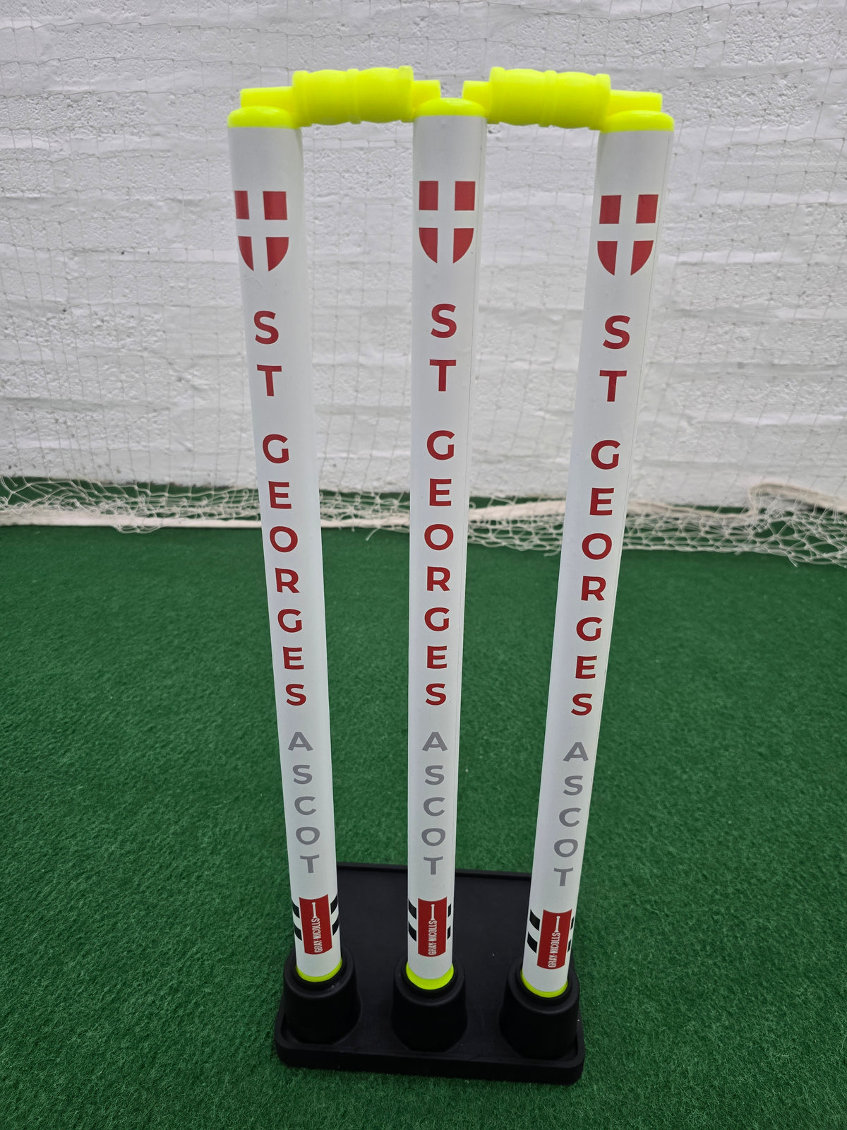 Three white Gray Nicolls Custom Printed Match Cricket Stumps with ST GEORGES ASCOT in red and grey on a heavy rubber base, topped with yellow bails, are set on green artificial turf against a netted background.