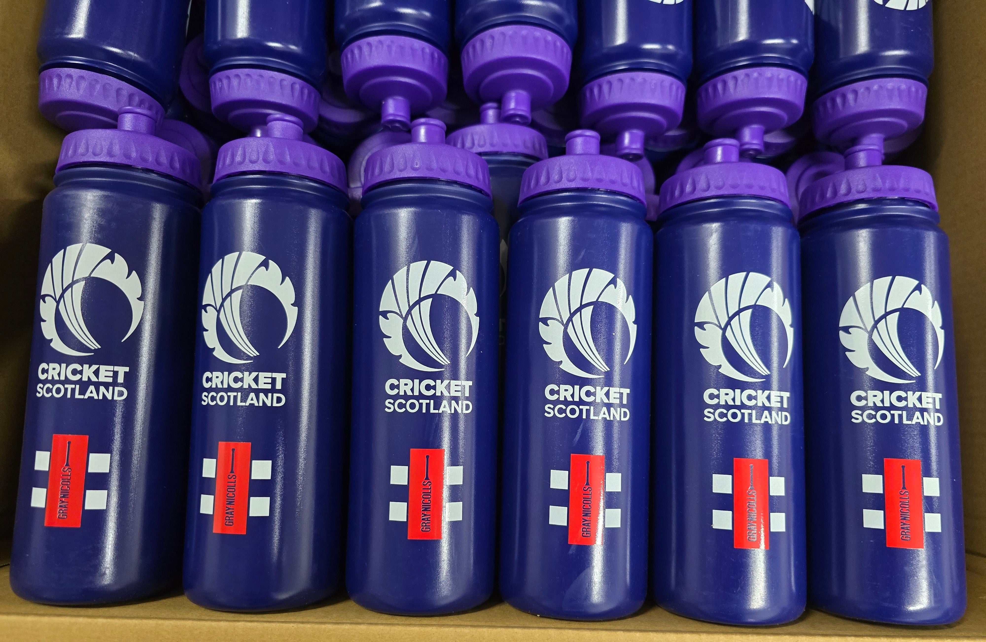 A close-up of multiple blue Martin Berrill Sports Customised Branded Water Bottles, each with a purple lid and club or school logo, neatly arranged in rows inside a cardboard box.