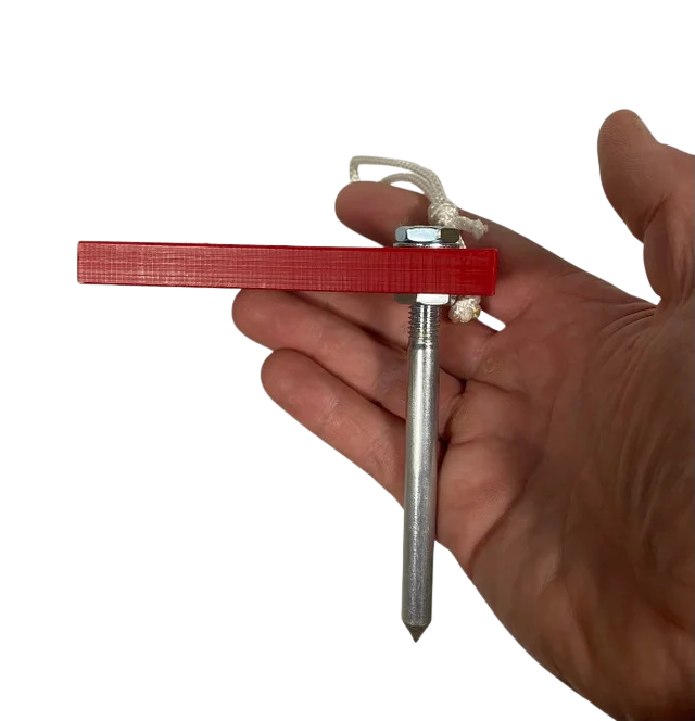A hand holds the Fusion Multi-Stump Spike HG by Fusion—a metal spike with a pointed end for hard ground, featuring a red rectangular plastic piece and a white cord tied to a nut at the top.