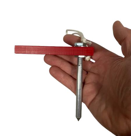 A hand holds the Fusion Multi-Stump Spike HG by Fusion—a metal spike with a pointed end for hard ground, featuring a red rectangular plastic piece and a white cord tied to a nut at the top.