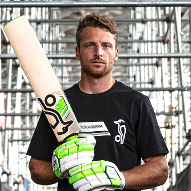 A man in a black Kookaburra t-shirt and green-and-white gloves holds a Kookaburra Kahuna Jos Buttler Pro Players Replica Cricket Bat 2026, standing before metal scaffolding and looking directly at the camera.