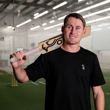 A man in a black t-shirt smiles while holding the Kookaburra Ghost Marnus Labuschagne Pro Players Replica Cricket Bat 2026 by Kookaburra over his shoulder inside an indoor cricket practice facility.