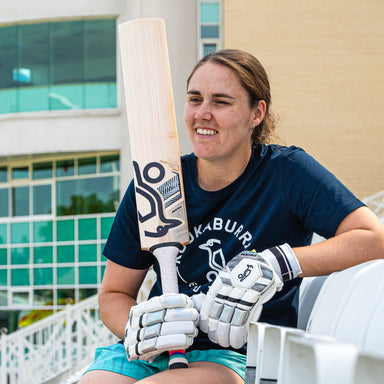 A smiling woman wearing cricket gloves sits on white steps outdoors, holding a Kookaburra Prism Natalie Sciver-Brunt Pro Players Replica Cricket Bat 2026 by Kookaburra, with a modern building in the background.