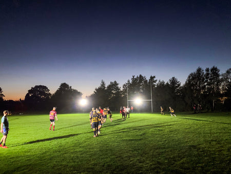 Rugby players compete at dusk on a grassy field, illuminated by Ritelite's Sports Lite Portable Floodlights, with trees and a darkening sky in the background.
