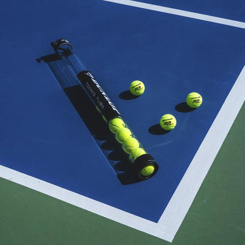 A Dunlop Pick-up Tube, partially filled with yellow tennis balls, rests on a blue and green tennis court with white lines. Three loose Dunlop tennis balls are scattered nearby.