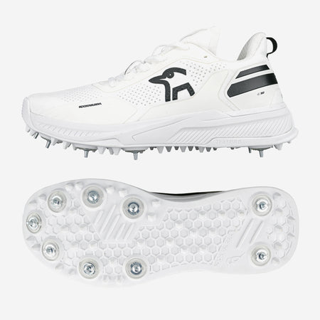 The Kookaburra KC Players Cricket Spikes are white shoes with black accents, a side logo, and a honeycomb-patterned sole featuring prominent metal spikes for grip. Shown from the side and bottom.