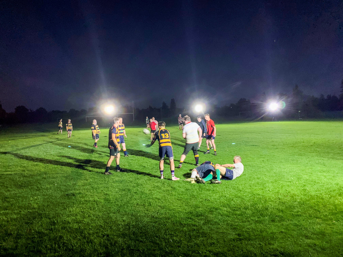 A group of rugby players practice at night on a field brightly lit by Ritelite Sports Lite Portable Floodlights. Some players chat while two are on the ground. Trees are faintly visible in the dark background.