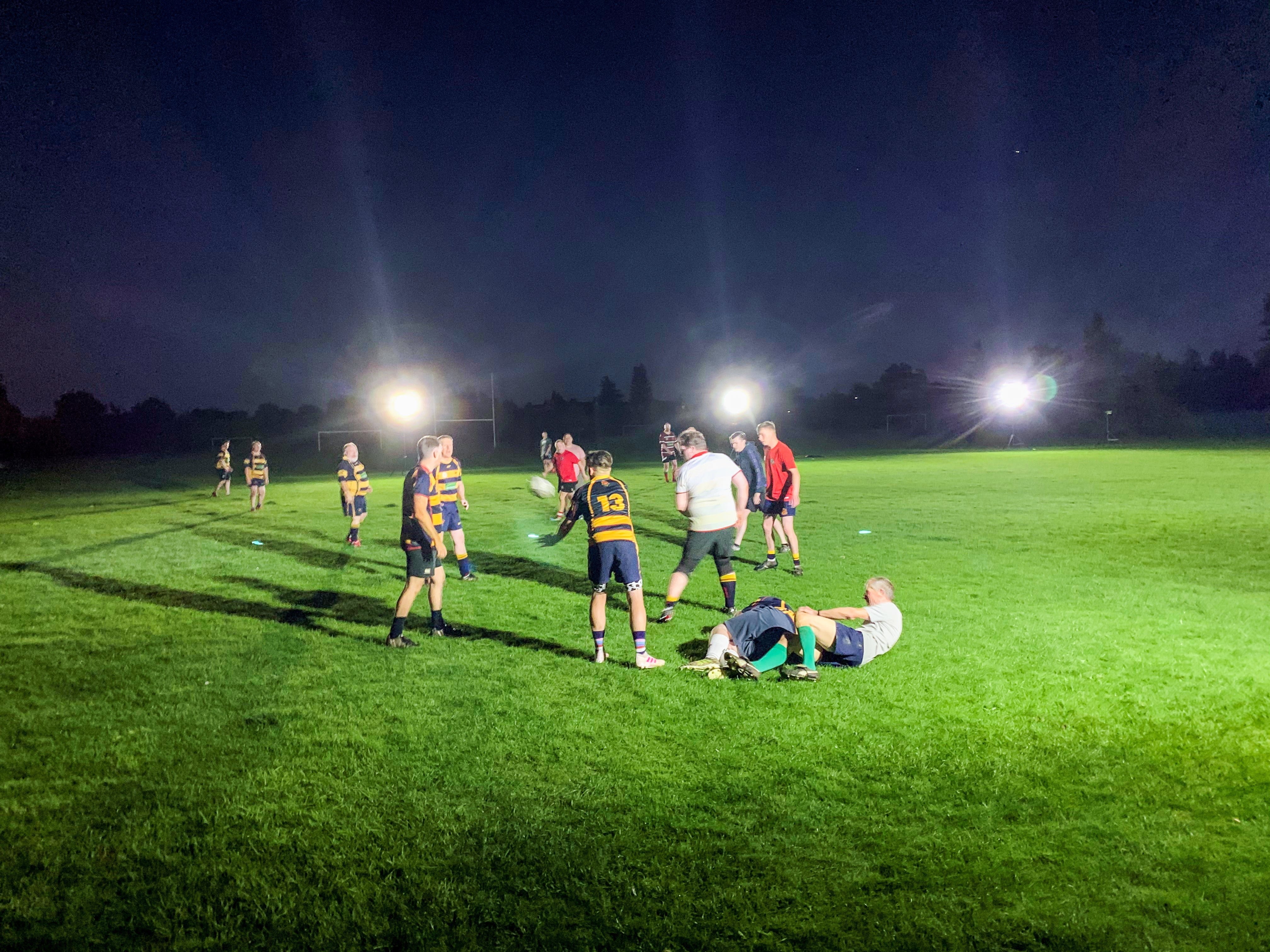 A group of rugby players practice at night on a field brightly lit by Ritelite Sports Lite Portable Floodlights. Some players chat while two are on the ground. Trees are faintly visible in the dark background.