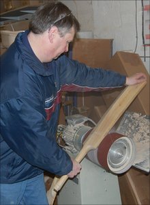 Wearing a blue jacket in a workshop with boxes and tools, a person uses the Martin Berrill Sports Cricket Bat Repair - Full Refurbishment service to restore a wooden cricket bat with a sanding machine.