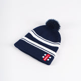The Gray Nicolls Bobble Hat is a navy knit hat with white horizontal stripes, a pom-pom on top, and a folded brim featuring a red rectangular patch with white text and a small graphic.