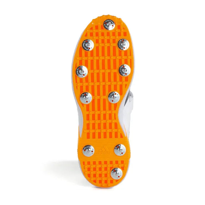 The image displays the sole of the Adidas 22YDS Vector White/Orange Cricket Spikes 2026 with a vibrant orange tread and 10 metal spikes for traction, making them ideal for pace bowlers.