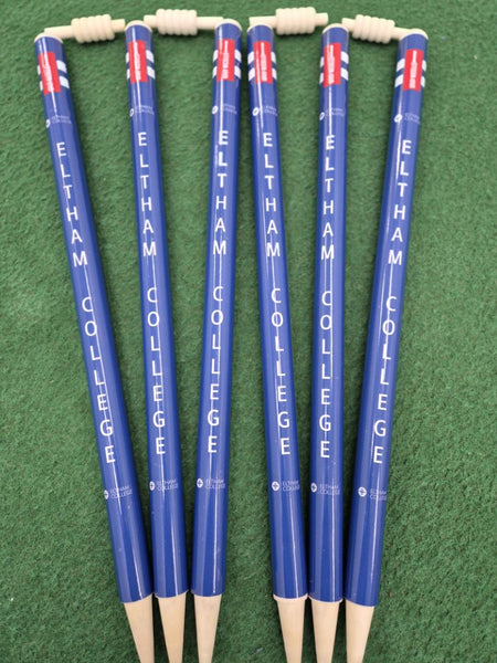 Six Gray Nicolls Custom Printed Club Cricket Stumps, blue with 'ELTHAM COLLEGE' printed in white vertically, are arranged side by side on green turf. Each stump features a white top secured by a coil.