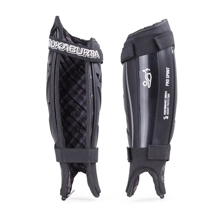 Pair of black Kookaburra Pro Spirit Shinguards: one side shows padded foam backing, the other reveals a hard shell with white logo. Both include ankle straps, foot loops, and reinforced spine for extra support.