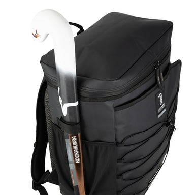 The Kookaburra Elite Hockey Rucksack 2026 is a black, heavy-duty backpack with zippered compartments, a laptop sleeve, and adjustable straps, shown holding a white and brown Kookaburra field hockey stick in its side pocket.