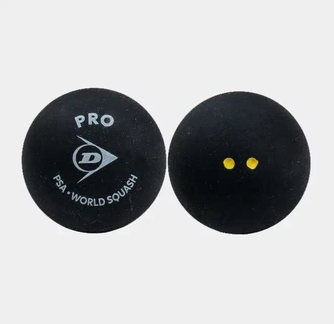 Set against a plain white background, two black Dunlop Pro Squash Balls (12 x 1 Ball Box) are shown. One ball features the “PRO PSA WORLD SQUASH” text and the Dunlop logo, while the other displays two small yellow dots—signifying its low bounce designed for professional-level play. Both balls have a matte texture, highlighting their premium quality from the trusted brand Dunlop.