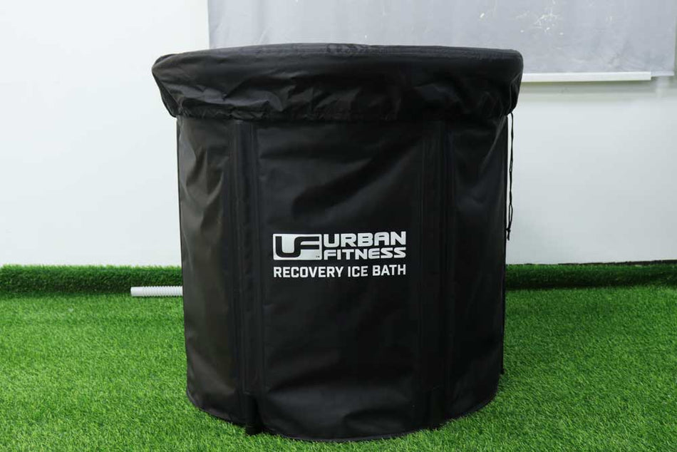 The Urban Fitness Recovery Ice Bath in black with a lid sits on artificial grass, perfect for cold therapy sessions, with a white wall and grey curtain as backdrop.