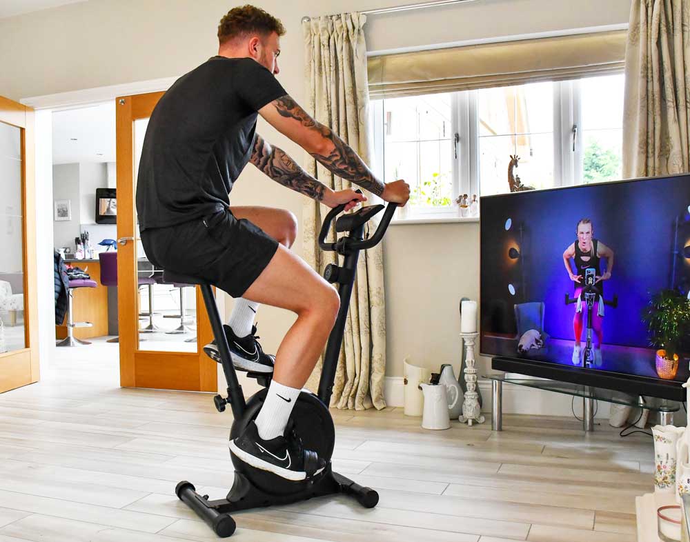 A man works out indoors on the Urban Fitness Magnetic Exercise Bike by Urban Fitness, adjusting resistance while following a virtual cycling class on TV in a bright room with wooden floors and large windows—ideal for home cardio workouts.