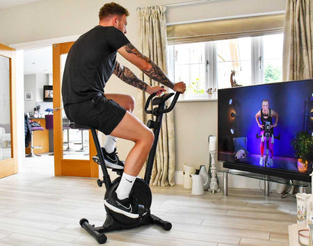 A man works out indoors on the Urban Fitness Magnetic Exercise Bike by Urban Fitness, adjusting resistance while following a virtual cycling class on TV in a bright room with wooden floors and large windows—ideal for home cardio workouts.