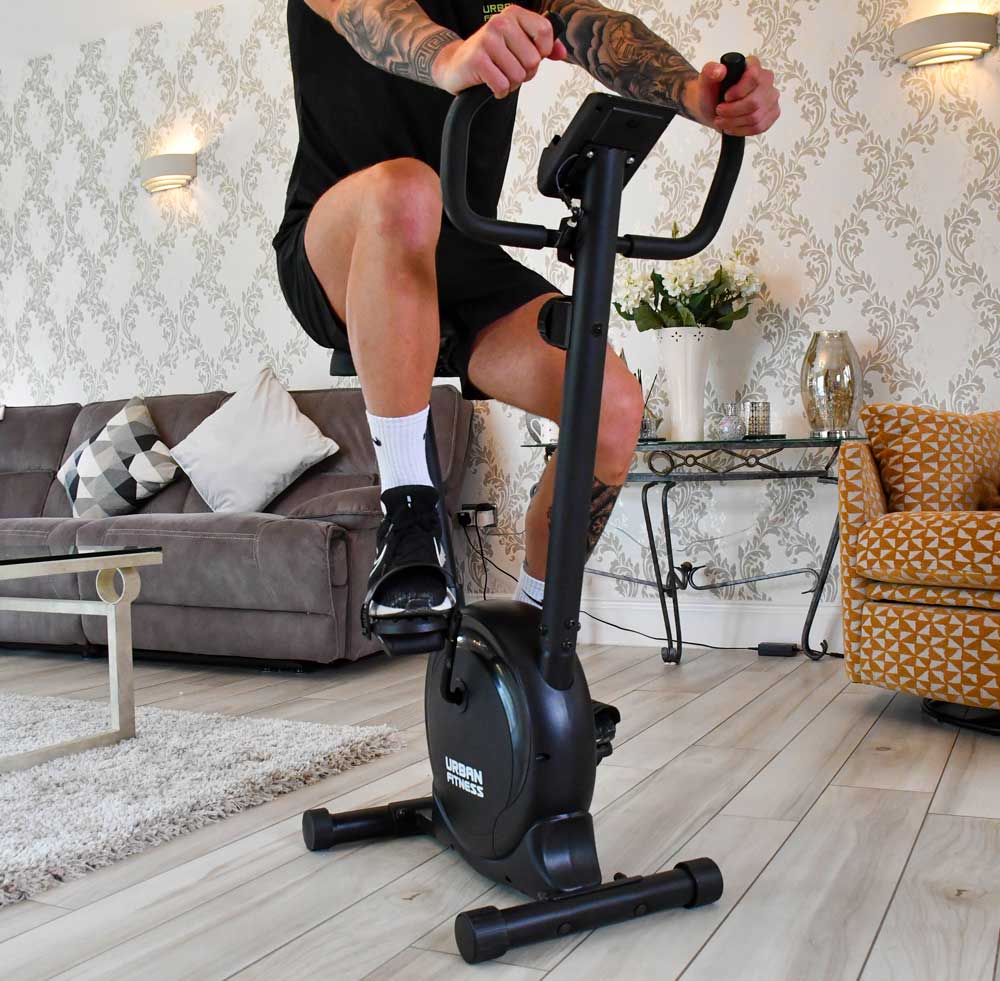 A person enjoys a home cardio session on the Urban Fitness Magnetic Exercise Bike by Urban Fitness, set in a stylish living room with a grey sofa, patterned wallpaper, and a yellow armchair.