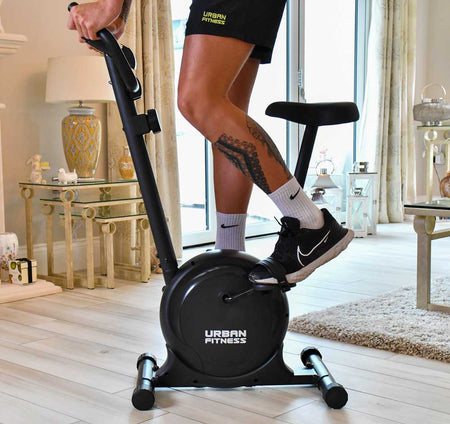 A person with a geometric leg tattoo uses the Urban Fitness Magnetic Exercise Bike by Urban Fitness for home cardio in a bright, modern living room featuring light wood floors and elegant decor.