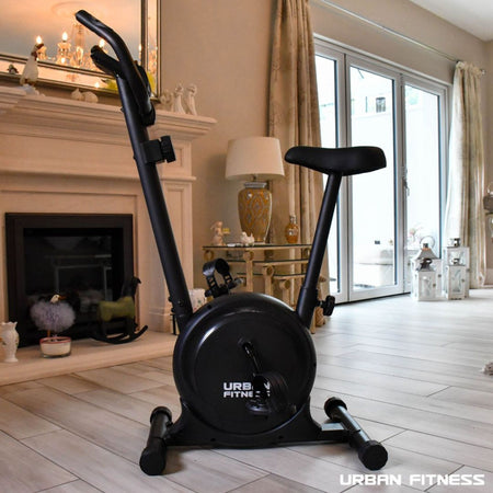 The Urban Fitness Magnetic Exercise Bike is set in a stylish living room with wooden floors, near large windows and a fireplace. The URBAN FITNESS logo appears in the corner—a sleek cardio solution for your home.