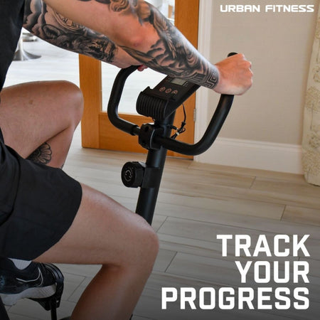 A person with tattooed arms pedals the Urban Fitness Magnetic Exercise Bike indoors. "Urban Fitness" and "Track Your Progress" appear, showcasing adjustable resistance for a personalized home cardio workout.