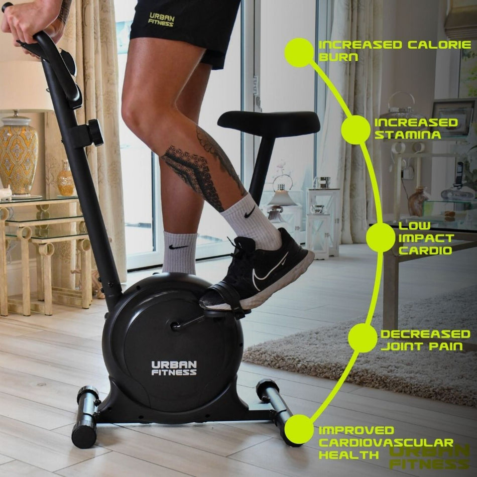 A person uses the Urban Fitness Magnetic Exercise Bike (Black) indoors, with labels showing benefits: higher calorie burn, more stamina, low-impact home cardio, less joint pain, better heart health, and adjustable resistance for custom workouts.