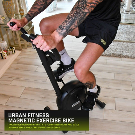 A person in black shorts and Nike shoes rides the Urban Fitness Magnetic Exercise Bike indoors. Text: Urban Fitness Magnetic Exercise Bike—ideal for home cardio, with adjustable resistance for customized intensity.