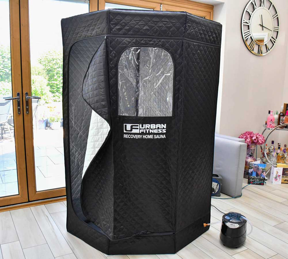 The Urban Fitness Recovery Home Sauna by Urban Fitness, in black, is set up near glass doors in a modern living room with light wood floors. Its transparent window and included steam generator offer relaxing heat therapy at home.