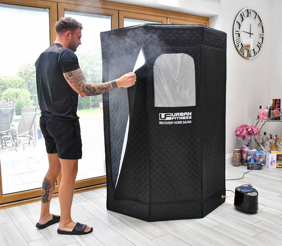 A man in sportswear unzips the Urban Fitness Recovery Home Sauna by Urban Fitness in a bright, modern room. A compact steam generator sits nearby, highlighting the convenience of home heat therapy.