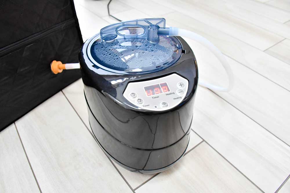 A close-up of the Urban Fitness Recovery Home Sauna steam generator by Urban Fitness, featuring a digital display, control buttons, clear lid with water droplets, and a white hose, sitting on a light-colored tile floor.