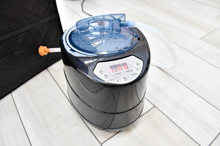 A close-up of the Urban Fitness Recovery Home Sauna steam generator by Urban Fitness, featuring a digital display, control buttons, clear lid with water droplets, and a white hose, sitting on a light-colored tile floor.