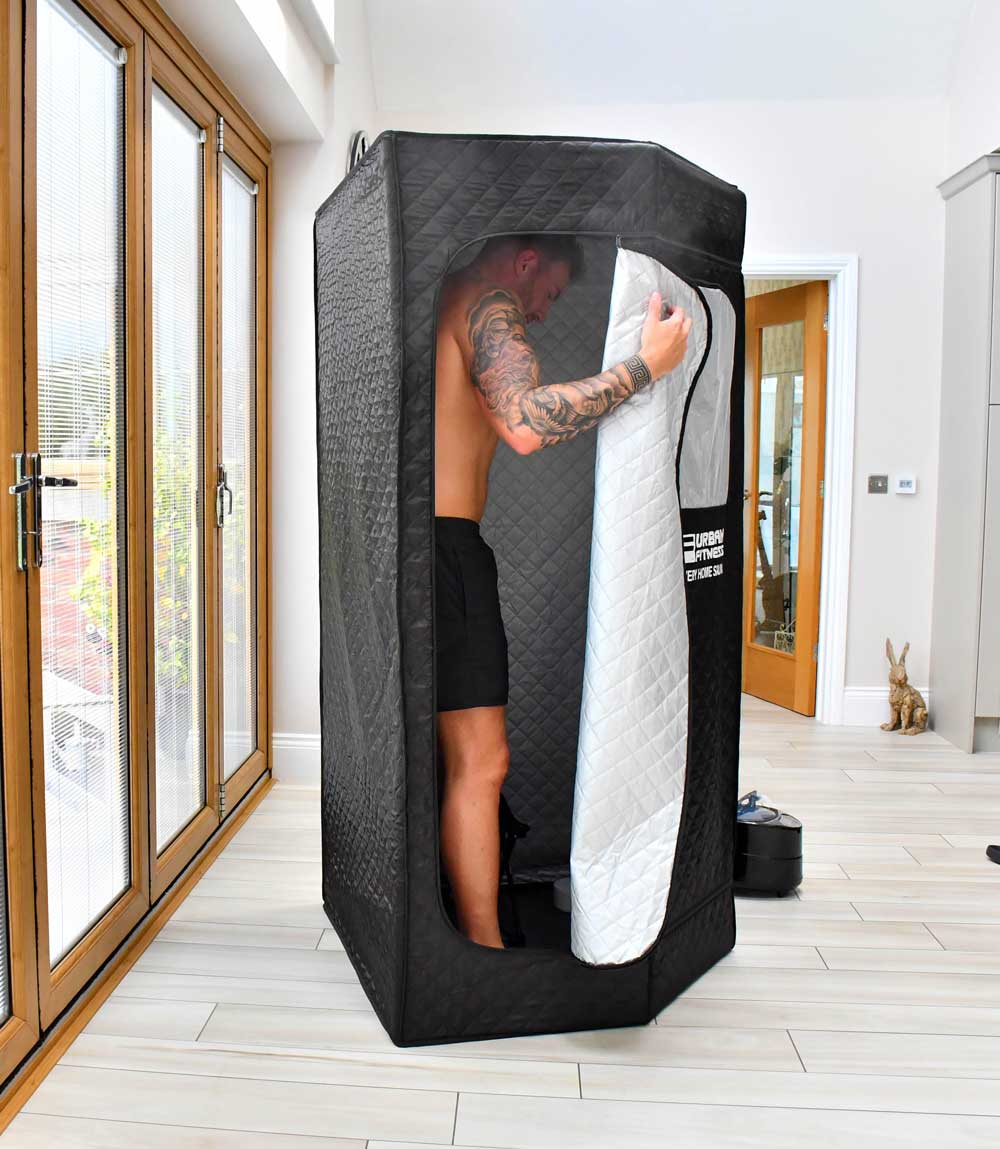 A man in shorts stands inside the Urban Fitness Recovery Home Sauna by Urban Fitness in a bright room with large windows, holding the tent's zippered door open. A small rabbit figurine is on the floor nearby.