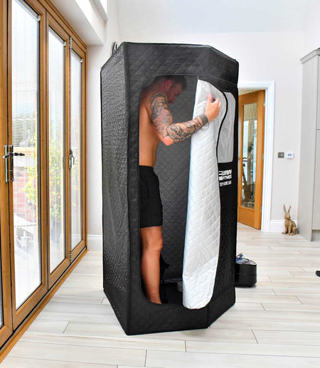 A man in shorts stands inside the Urban Fitness Recovery Home Sauna by Urban Fitness in a bright room with large windows, holding the tent's zippered door open. A small rabbit figurine is on the floor nearby.
