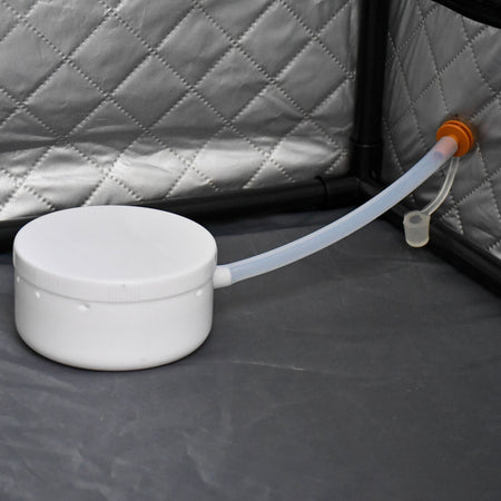 The Urban Fitness Recovery Home Sauna by Urban Fitness features a white plastic container with lid, connected via a flexible tube to an orange port on a quilted gray wall—perfect for convenient heat therapy at home.