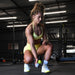 A woman in a yellow sports outfit and white sneakers squats indoors at the gym, holding a dumbbell and focusing on her workout with Urban Fitness Fitness Dice by Urban Fitness. Gym equipment can be seen in the background.