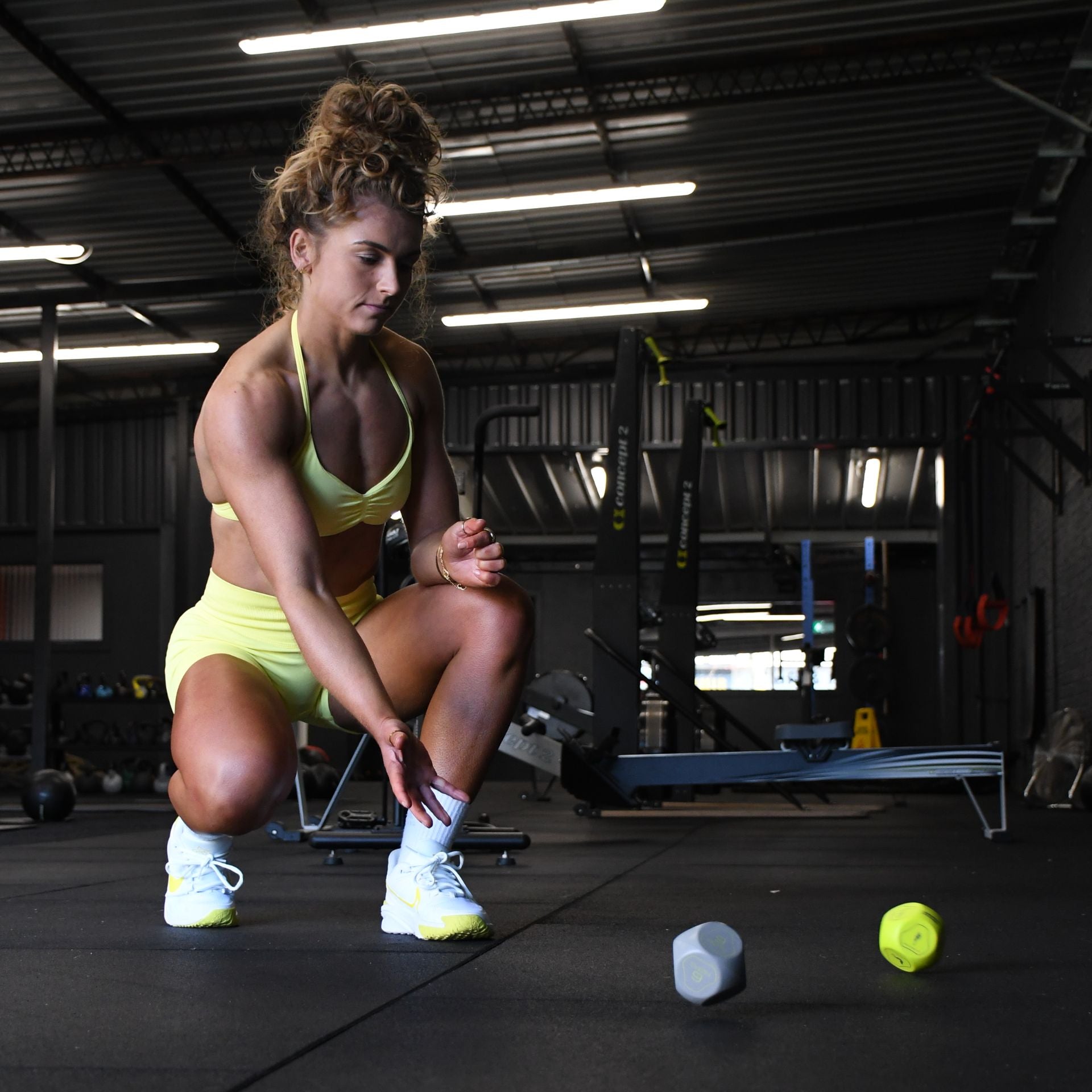 A woman in a yellow sports outfit kneels on the gym floor, watching two Urban Fitness Fitness Dice by Urban Fitness roll in front of her, with gym equipment visible in the background.