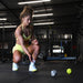 A woman in a yellow sports outfit kneels on the gym floor, watching two Urban Fitness Fitness Dice by Urban Fitness roll in front of her, with gym equipment visible in the background.