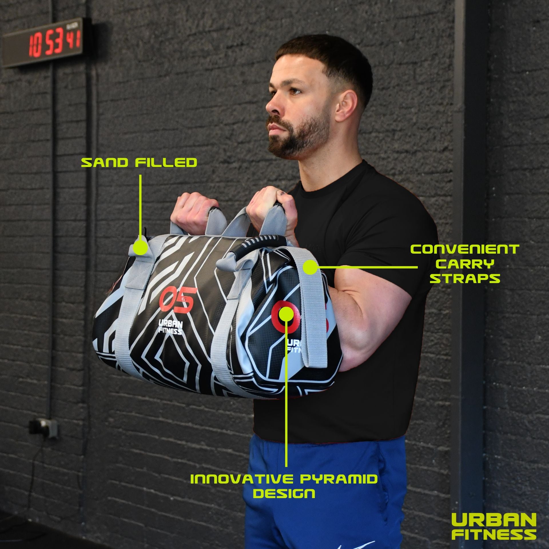 A man in a gym holds the Urban Fitness Power Bag by its convenient straps. Labeled features highlight its sand filling, functional design for full-body conditioning and strength. The Urban Fitness logo is visible in the corner.