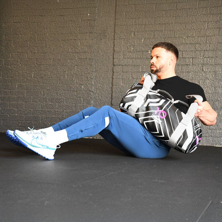 A man in blue pants and a black shirt does Russian twists with an Urban Fitness Power Bag, building functional strength in a gym with black walls and flooring.