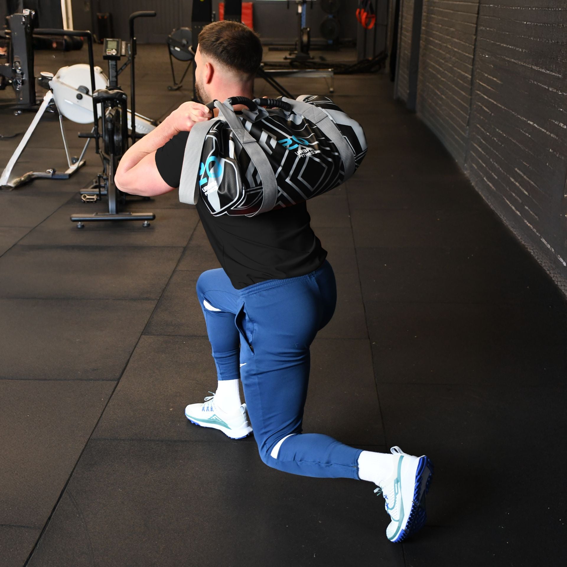 A man in a black shirt and blue pants lunges in the gym, holding an Urban Fitness Power Bag by Urban Fitness across his shoulders. Gym equipment and machines are visible in the background.