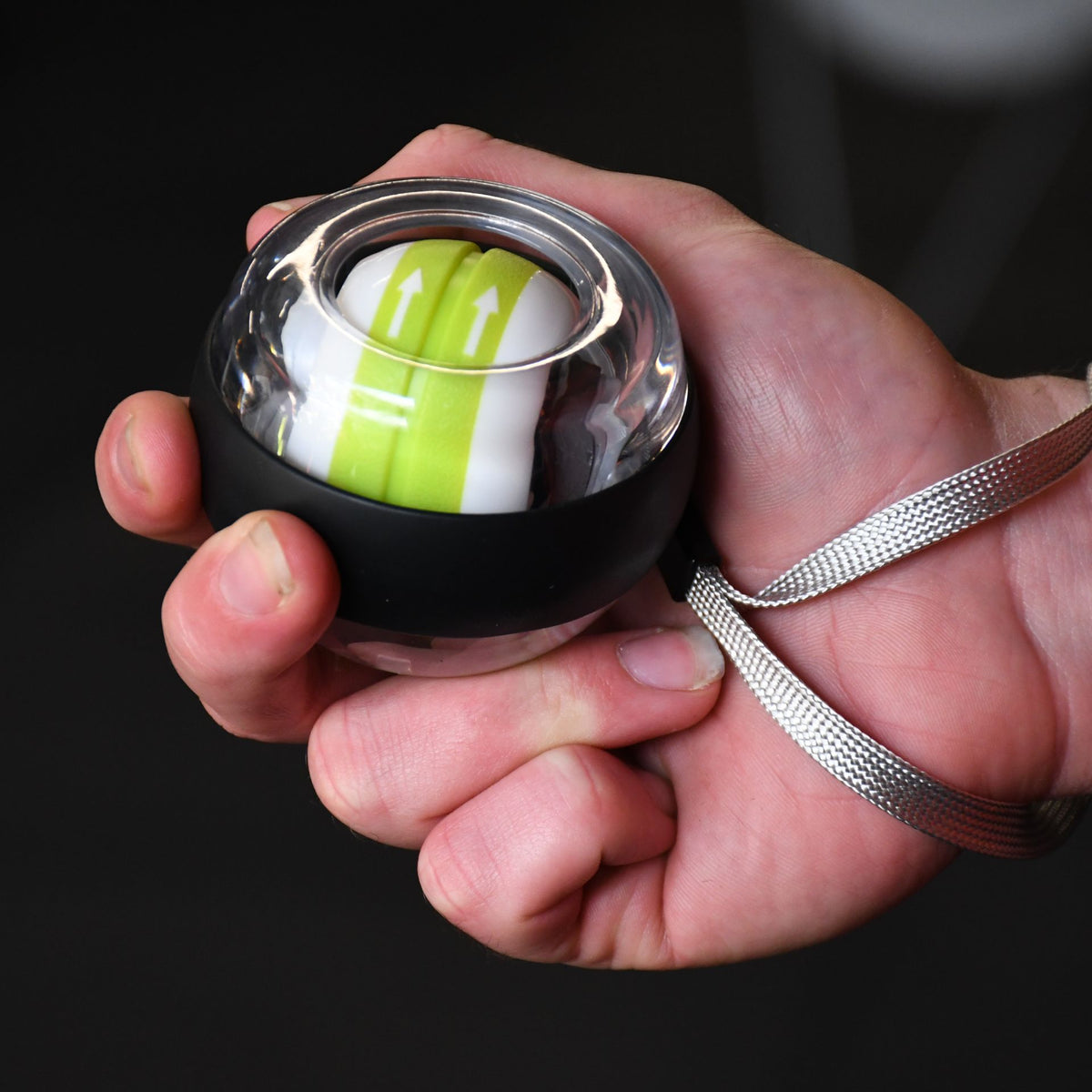 A person holds the Urban Fitness Wrist Exerciser Ball by Urban Fitness, featuring a small circular design with a black and transparent casing, green and white inner rotor, and silver wrist strap—ideal for grip strength and injury prevention.