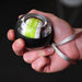 A person holds the Urban Fitness Wrist Exerciser Ball by Urban Fitness, featuring a small circular design with a black and transparent casing, green and white inner rotor, and silver wrist strap—ideal for grip strength and injury prevention.