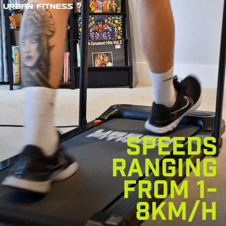 A person with a leg tattoo runs indoors on the Urban Fitness Walking Treadmill by Urban Fitness (speeds: 1–8km/h). Shelves behind display books and a record, highlighting the treadmill’s smart fitness tracking features.