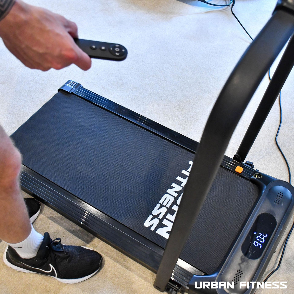 A person holding a remote control next to the Urban Fitness Walking Treadmill by Urban Fitness, ready for convenient workouts with smart fitness tracking.